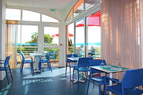 An indoor dining area with blue chairs and tables, large windows, and red umbrellas outside.. Residence Bella Vista