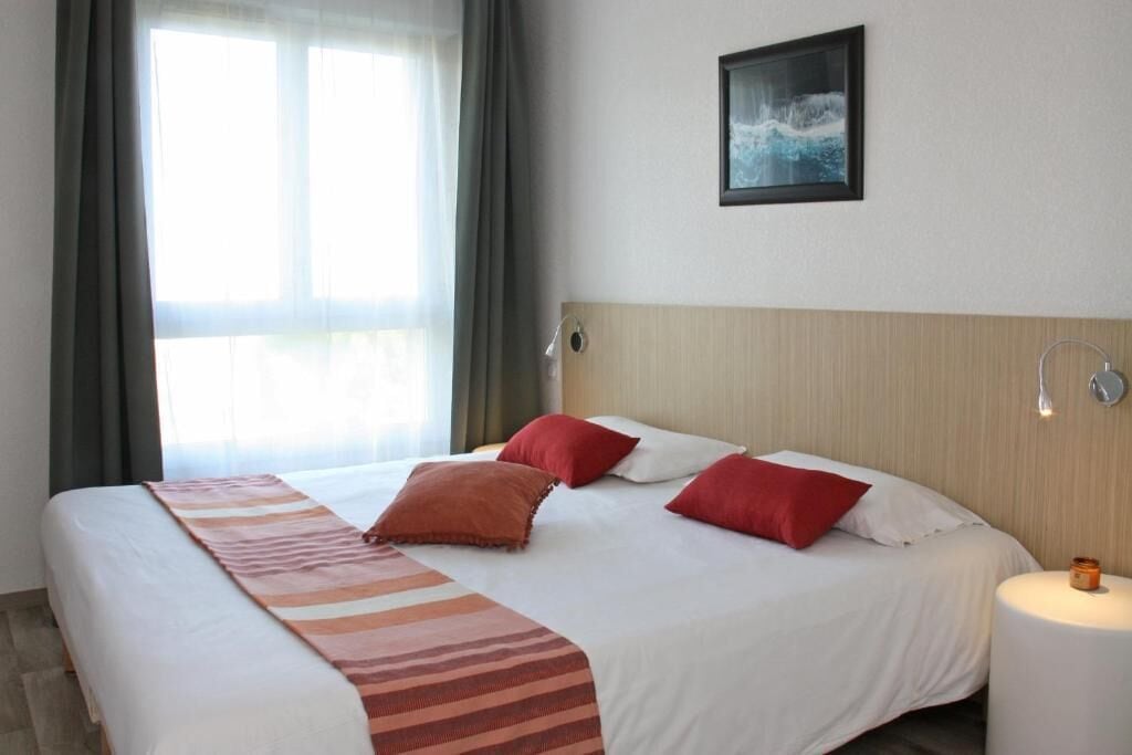 Habitación de hotel con una cama grande, cabecera, mesita de noche con lámpara y ventana con cortinas.