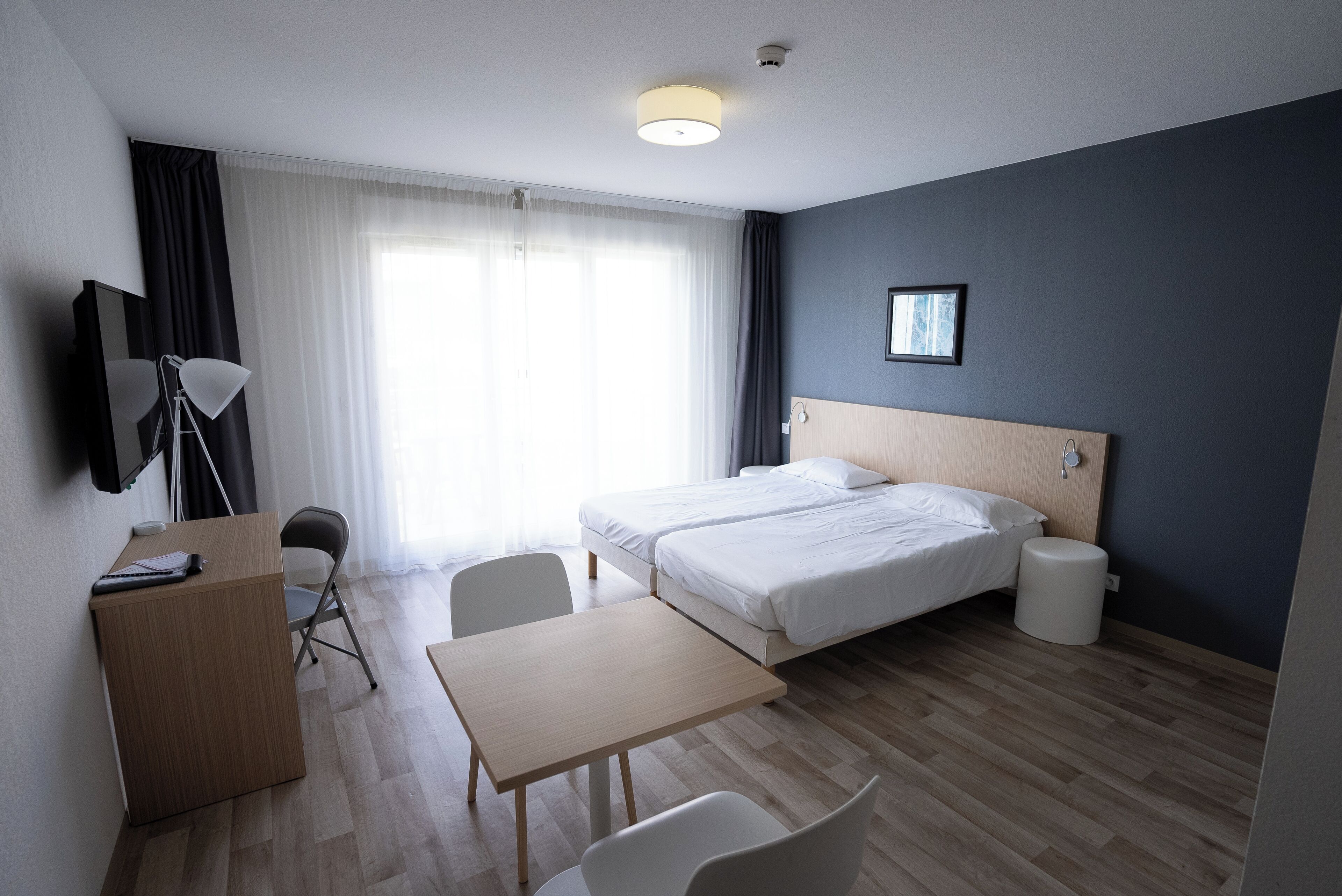 A hotel room with two beds, a desk with a chair, a TV, and a window with curtains.