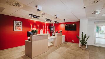Zona de recepción con pared roja, mostrador de recepción, televisor y planta en maceta.