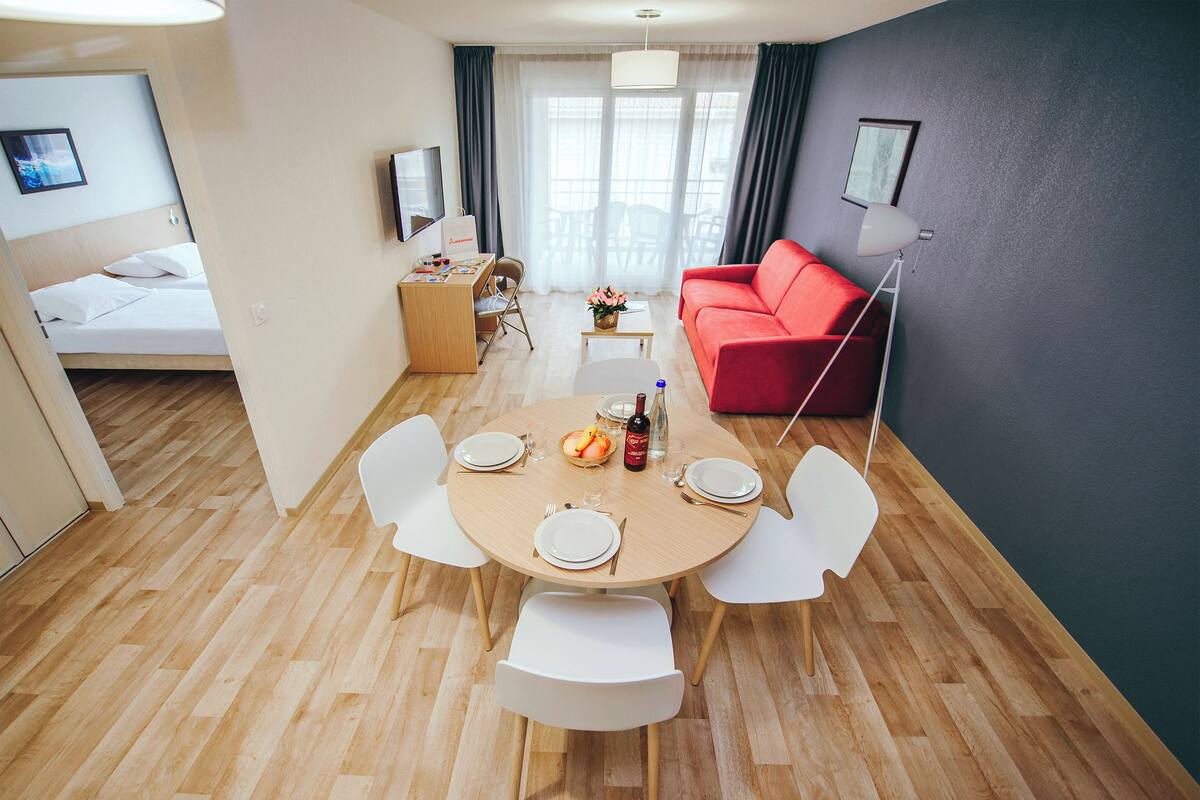 Un salon moderne avec un canapé rouge, une table à manger avec des chaises, un petit bureau et un lit à l’arrière-plan.