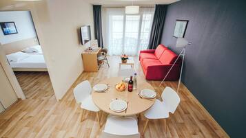 A modern living room with a red sofa, a dining table with chairs, a small desk, and a bed in the background.