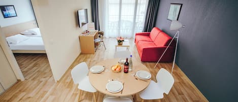 A modern living room with a red sofa, a dining table with chairs, a small desk, and a bed in the background.