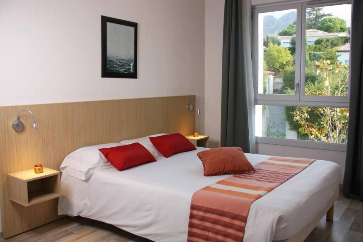 Une chambre d’hôtel avec un grand lit, deux oreillers rouges, une tête de lit en bois, une table de chevet avec une lampe et une fenêtre donnant sur un paysage verdoyant et des bâtiments.