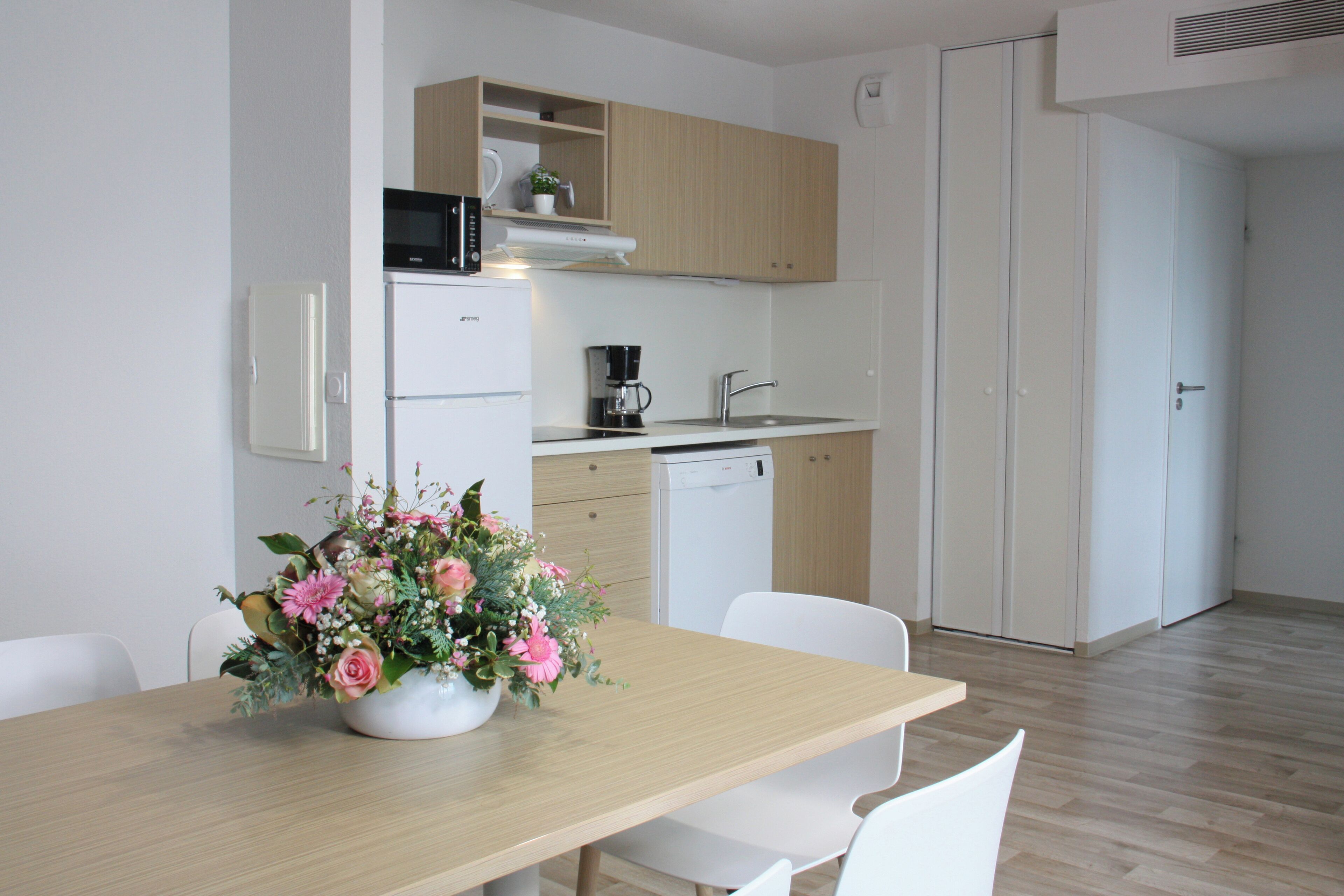 A modern kitchen with a microwave, coffee maker, and dishwasher. There is a dining table with chairs and a vase of flowers.