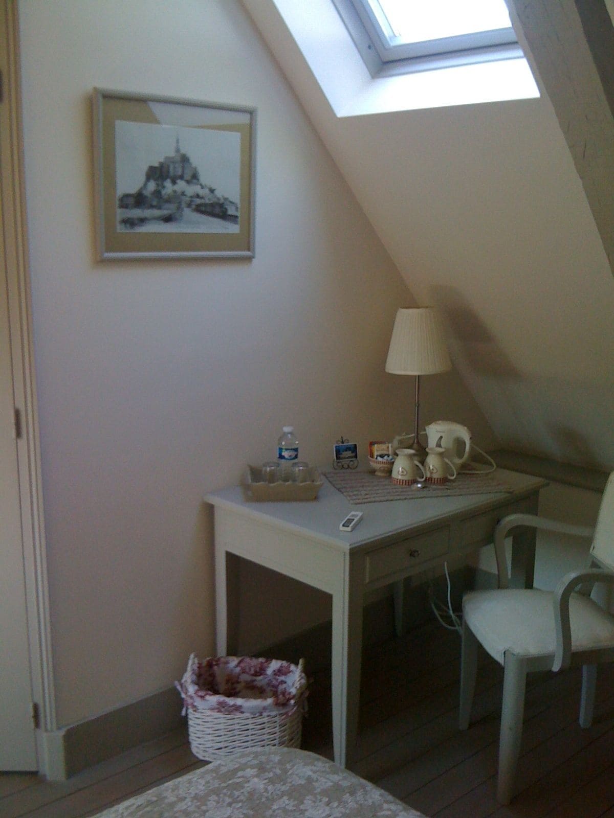 A small, cozy room with a sloped ceiling, a desk with a lamp, a chair, and a framed picture on the wall.