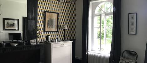Una habitación con pared de diseño blanco y negro, una mesa blanca con un letrero, una silla negra con cojín amarillo, una silla negra con diseño, un mueble negro para televisión y una ventana con cortinas.