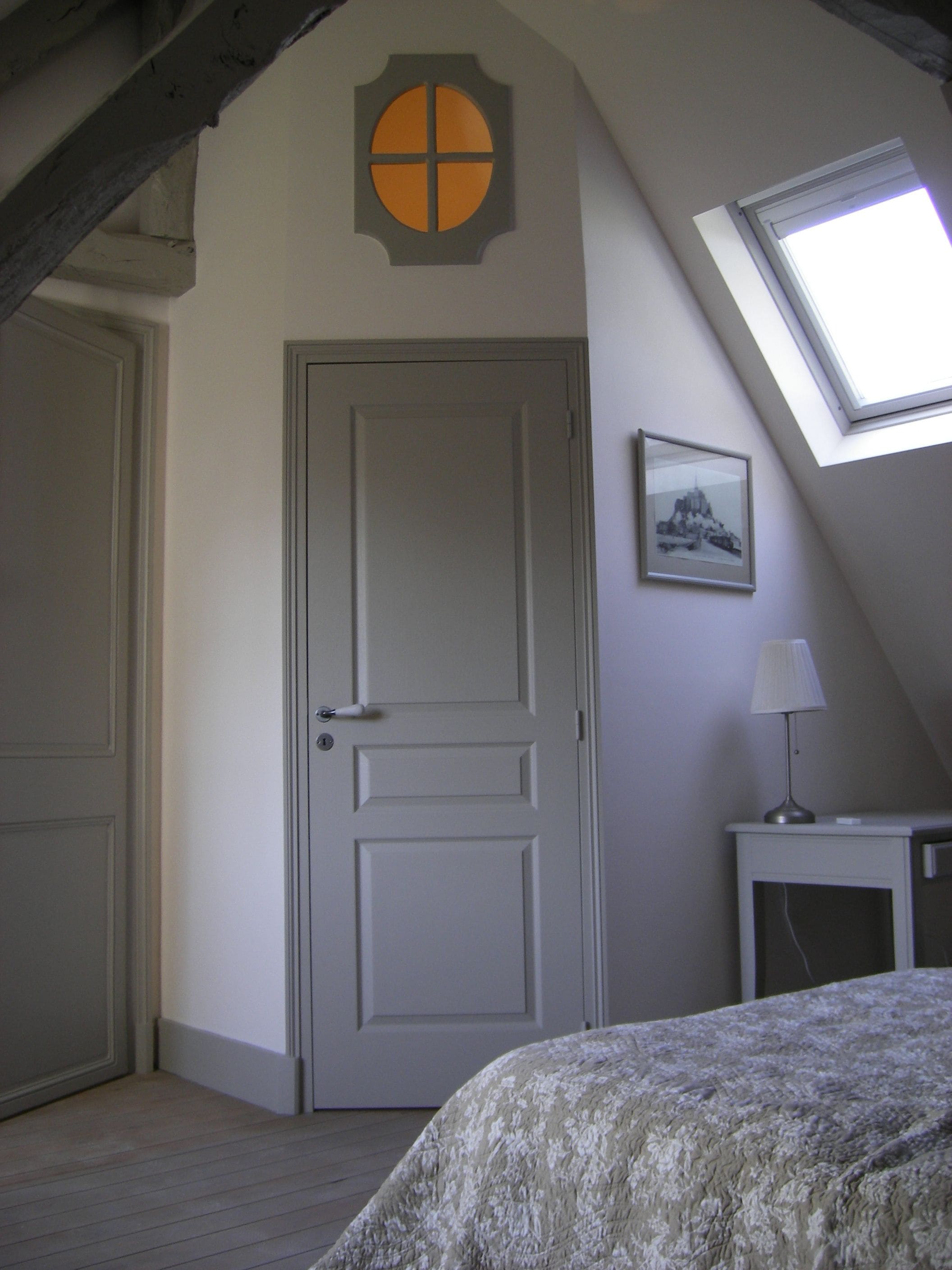 A bedroom with a sloped ceiling, a door, a small bedside table with a lamp, and a framed picture on the wall.