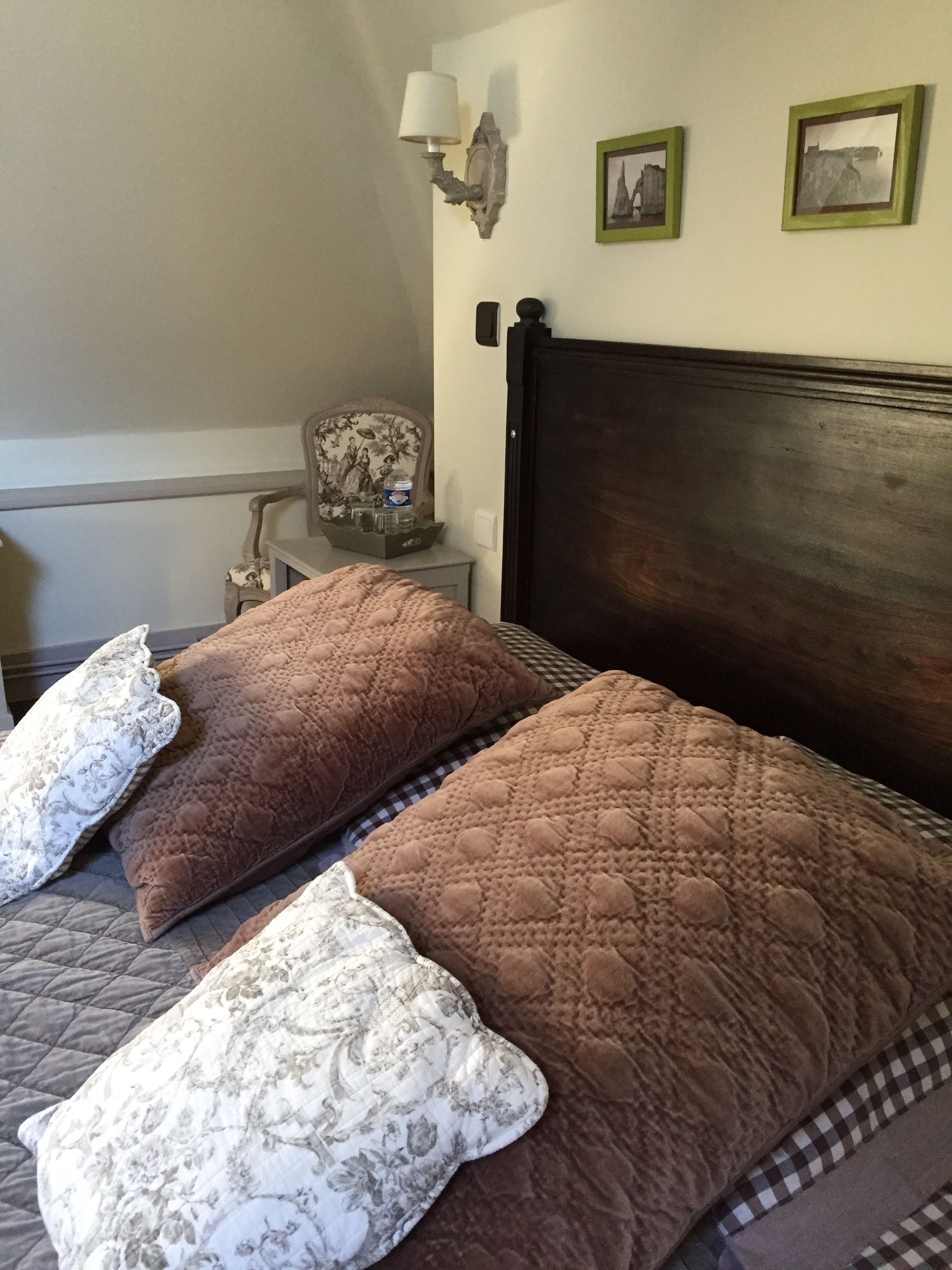 A bedroom with two beds, each with a brown quilted bedspread and a floral pillow. There is a nightstand with a lamp, a framed picture, and a small table with a decorative item.