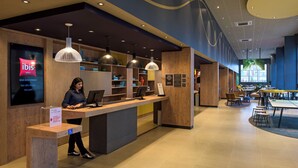 Zona de recepción de un hotel con un mostrador de madera, una computadora y una pantalla digital que muestra información del hotel. Se observan estanterías con diversos objetos y un área de descanso al fondo.