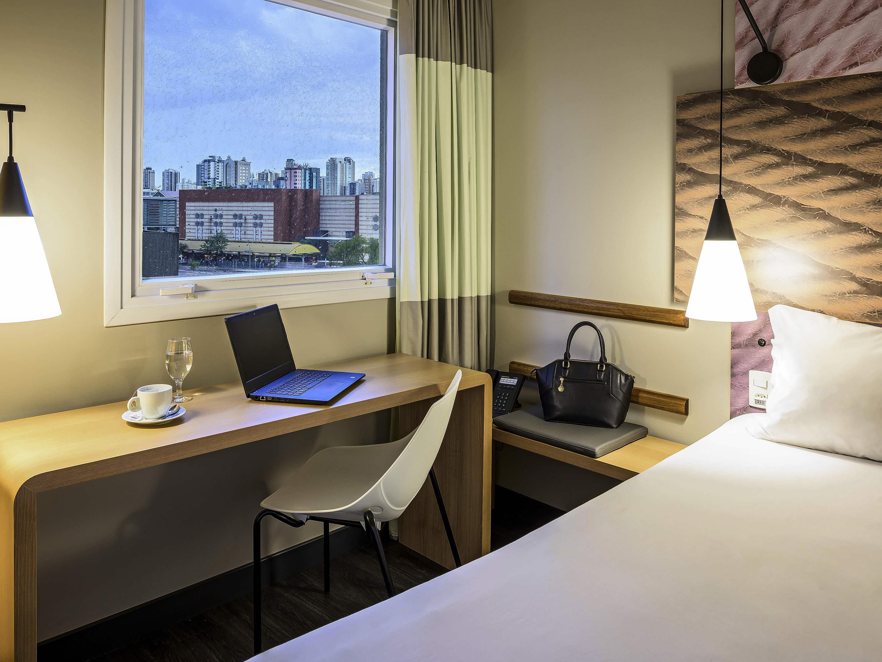 A hotel room with a desk, chair, laptop, bedside table with a handbag, and a bed.
