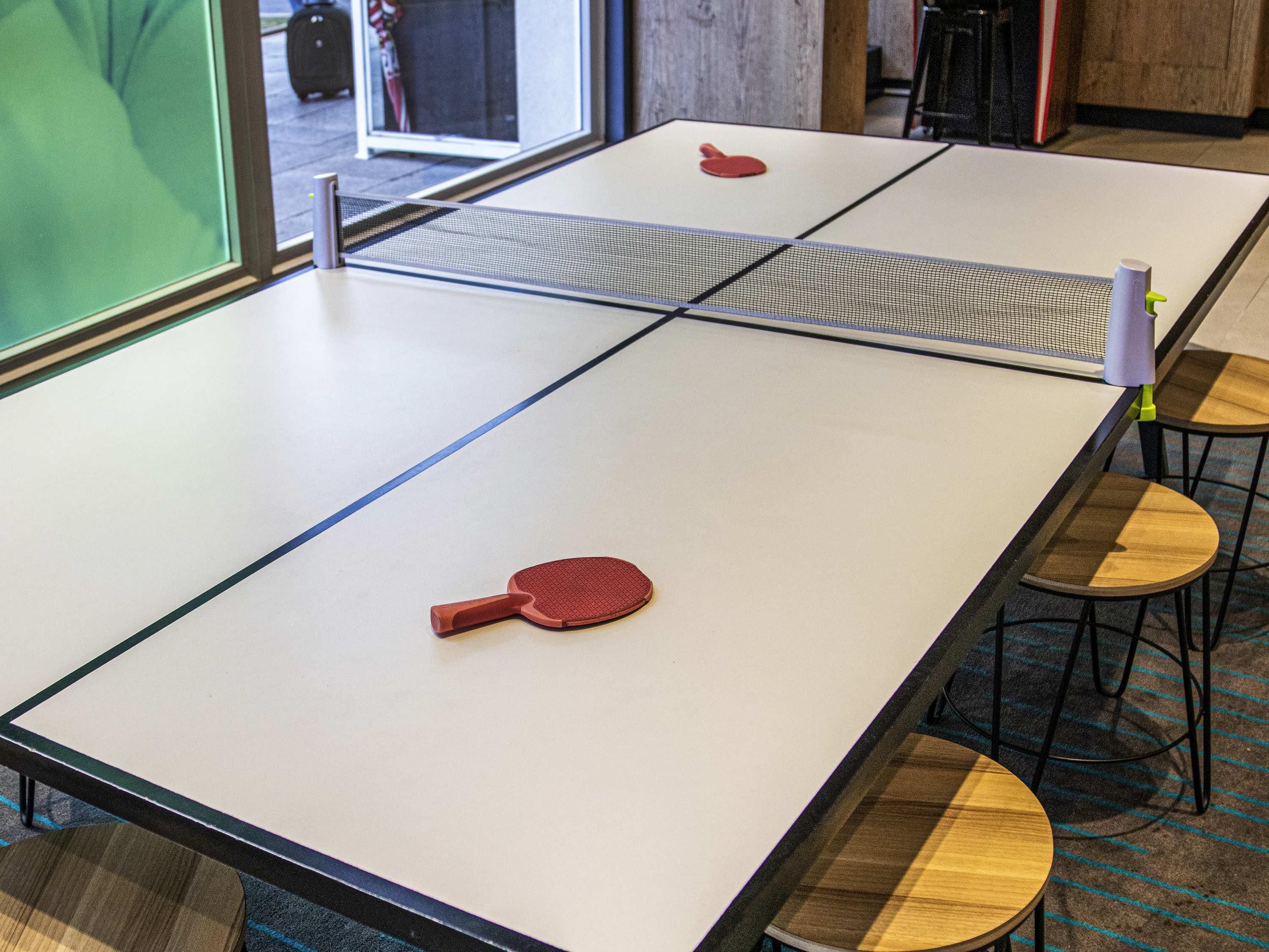 Une table de ping-pong avec deux raquettes et un filet au milieu. Il y a deux tabourets en bois avec des pieds en métal.