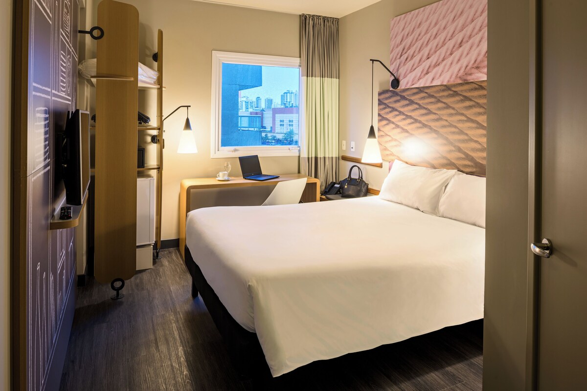 Une chambre d’hôtel comprenant un lit, un bureau avec un ordinateur portable, une fenêtre donnant sur la ville et une armoire.