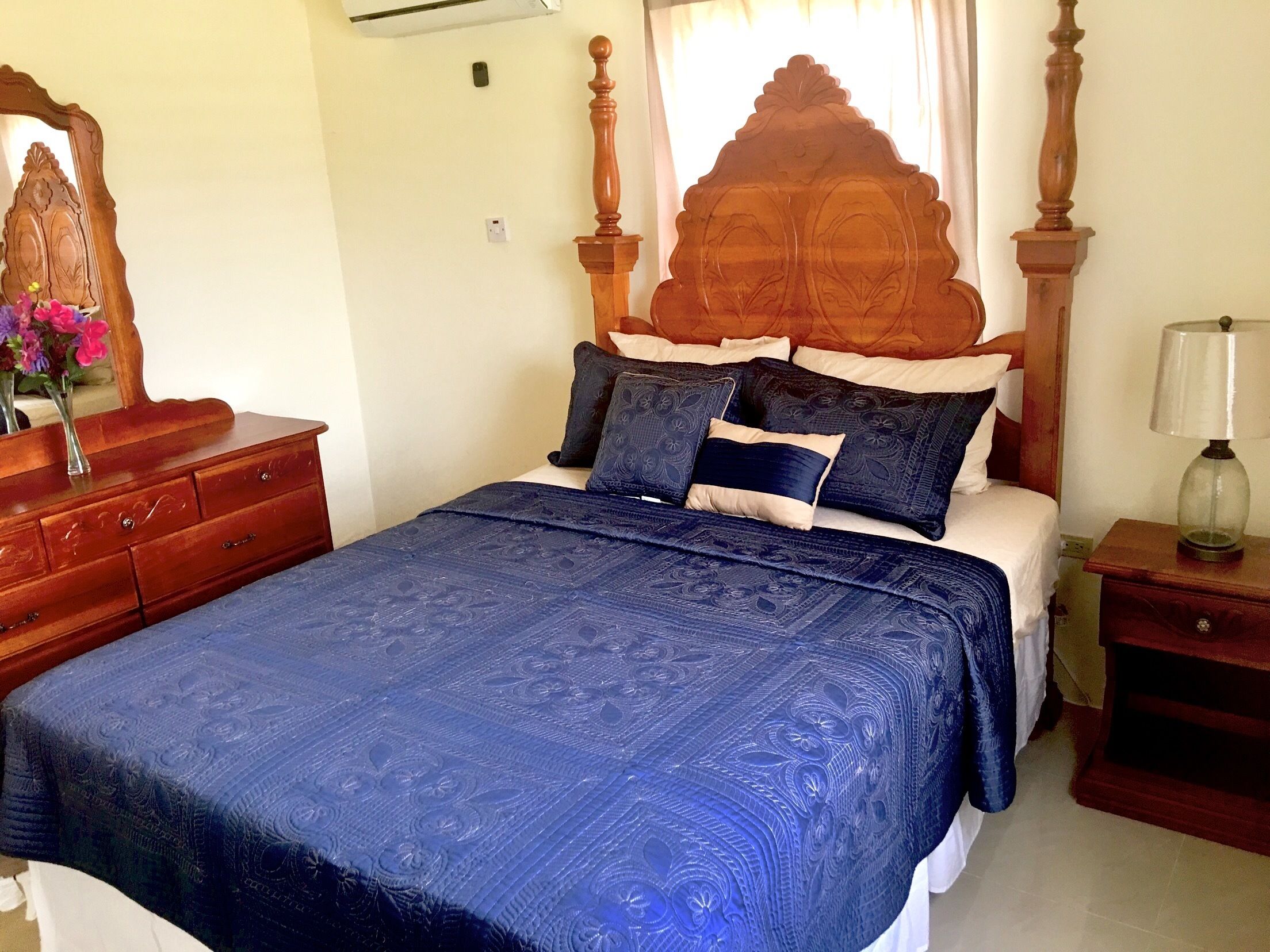 A bedroom with a wooden headboard, a bed with blue bedding, a nightstand with a lamp, and a dresser with a mirror and flowers.