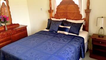 A bedroom with a wooden headboard, a bed with blue bedding, a nightstand with a lamp, and a dresser with a mirror and flowers.