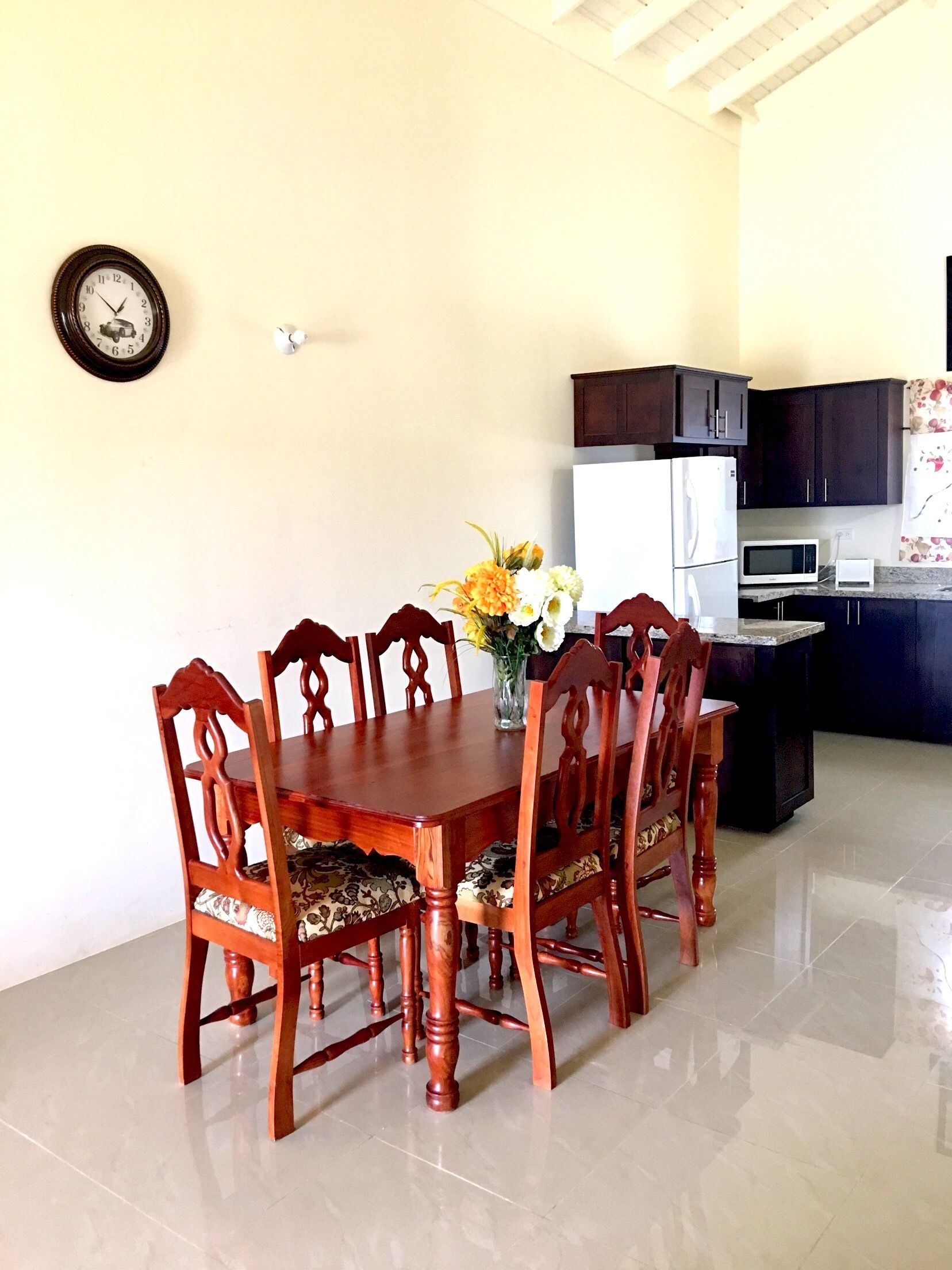 A dining area with a wooden table and chairs, a vase of flowers, a clock on the wall, and a kitchen in the background.