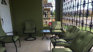 A patio area with four green chairs and a small table, a white door, and a view through a metal gate to a residential street. - Drax Hall Country Club's Sweet Escape (Ocho Rios)