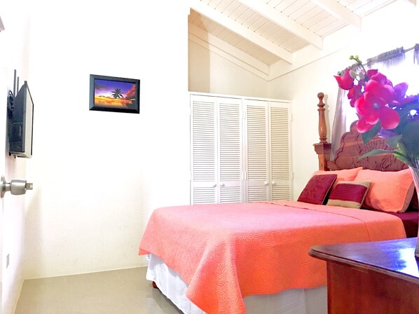 Une chambre à coucher avec un lit, une table de chevet, une tête de lit en bois, une armoire blanche à volets, un tableau au mur et une fenêtre donnant sur des palmiers.