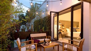 An outdoor patio with wooden furniture, a table with a bottle and glasses, and a view into a room with a bed and a nightstand. - H21 Hospedaje Boutique (Mexico City)