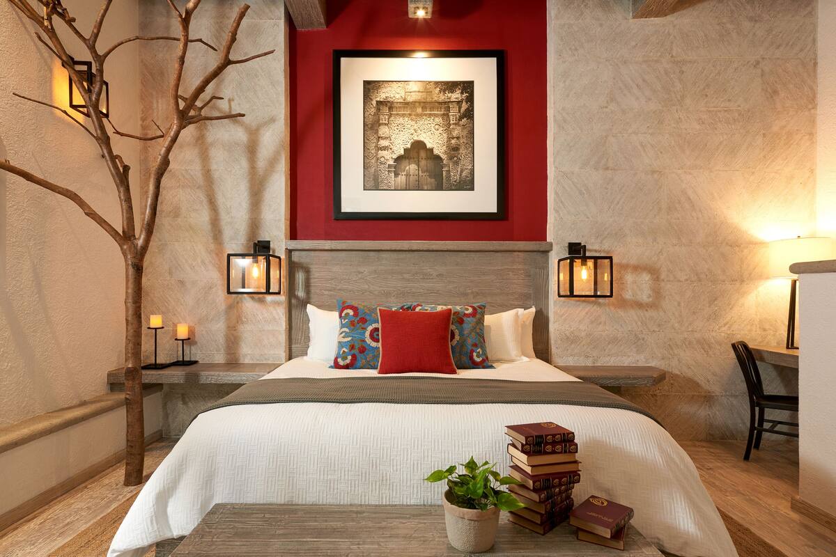 A bedroom with a wooden headboard, a framed picture above the bed, a bedside table with a lamp, and a bench with books and a plant.
