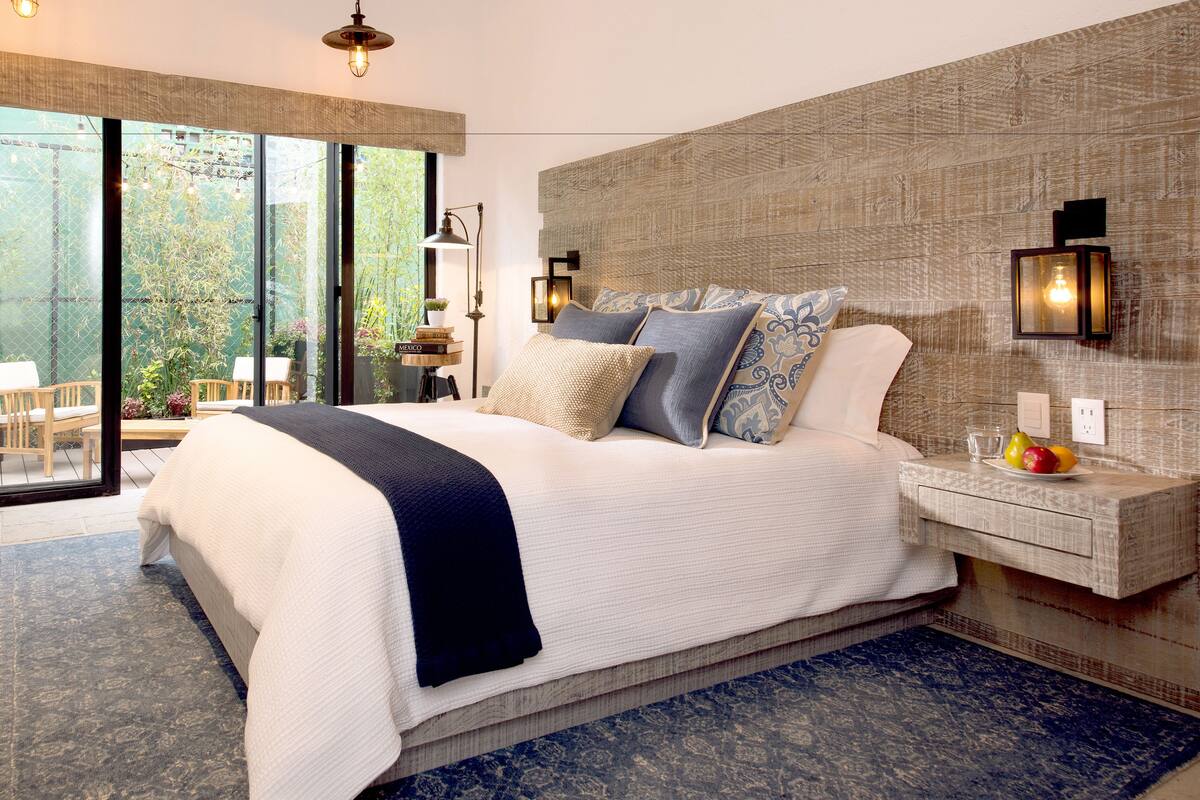 A modern bedroom with a large bed, a nightstand with a fruit bowl, and a view of greenery through the window.