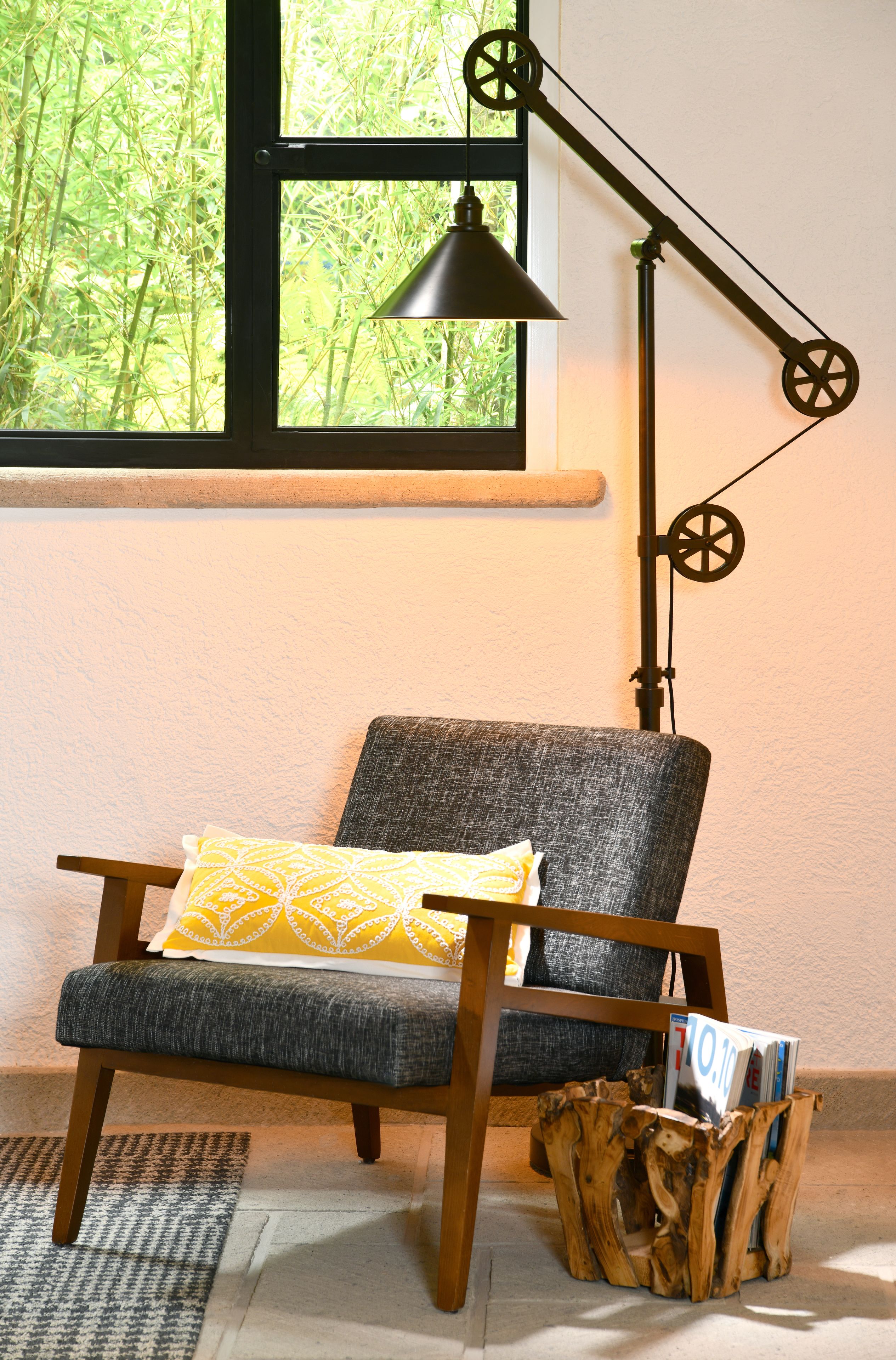 A chair with a cushion, a wooden floor, a window with greenery outside, and a floor lamp.