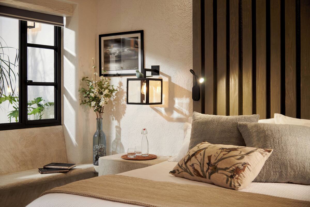 A cozy bedroom with a bed, a bedside table with a vase and books, a window with plants, and a wall-mounted lamp.