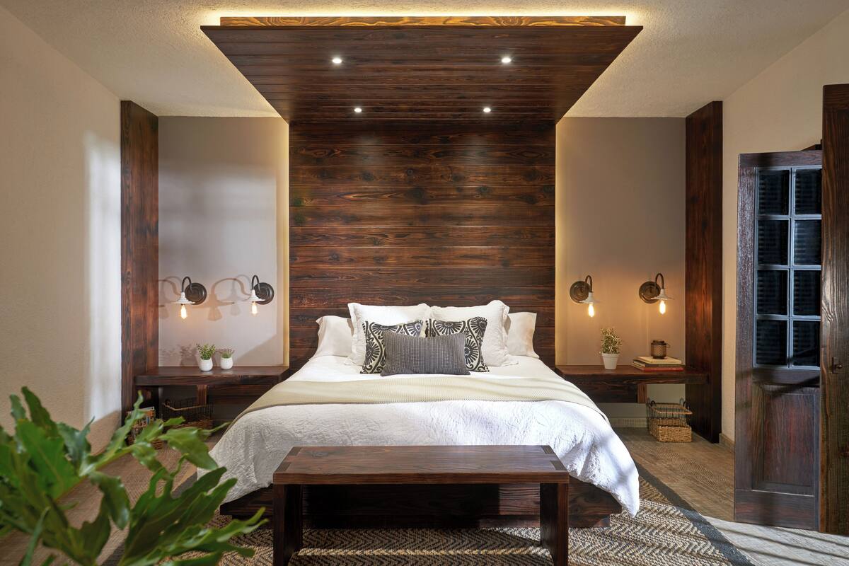 A bedroom with a wooden ceiling, a bed with white bedding, two bedside tables with lamps, and a small bedside plant.