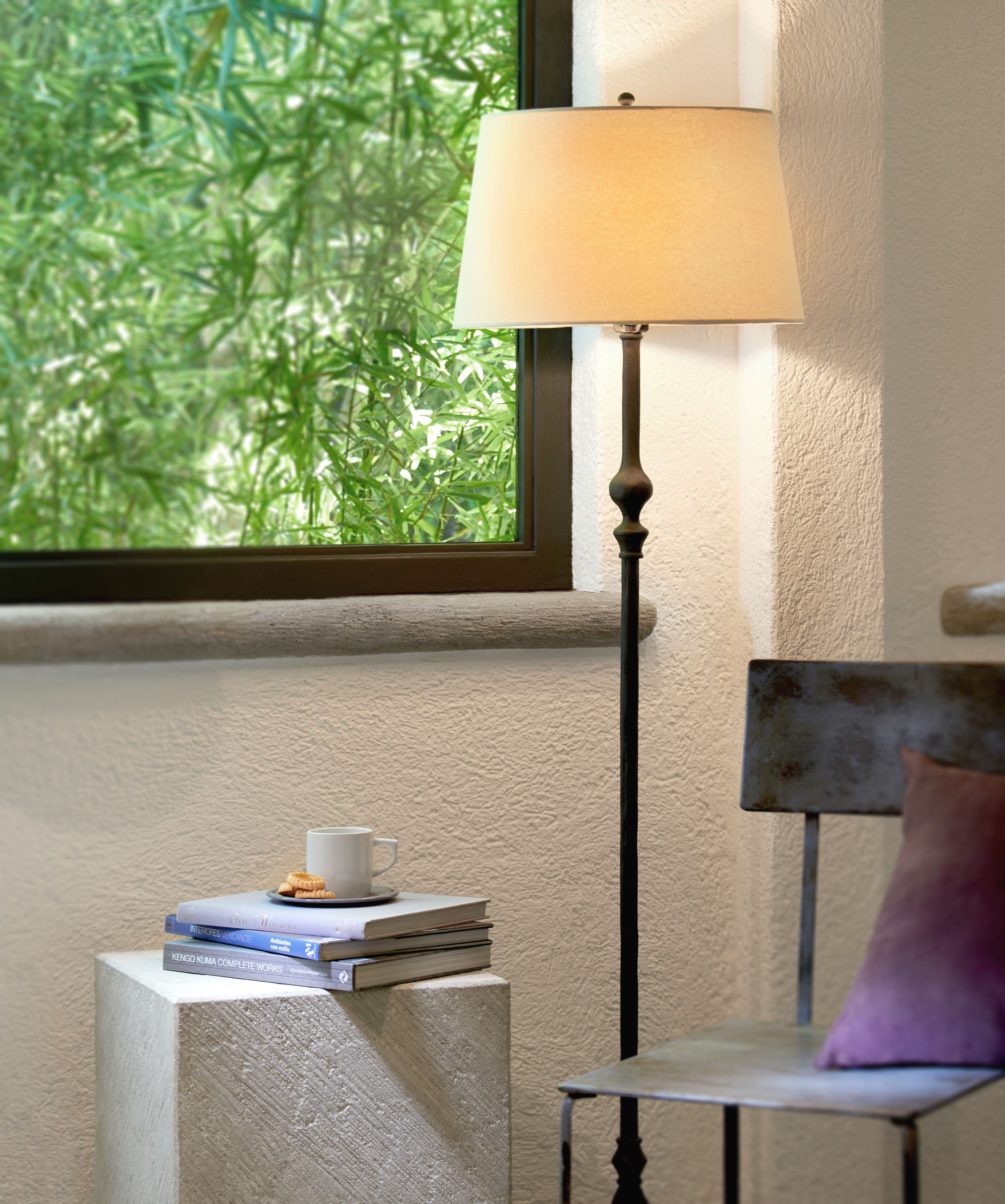 Una habitación con ventana, lámpara, silla y una pequeña mesa con libros y una taza.
