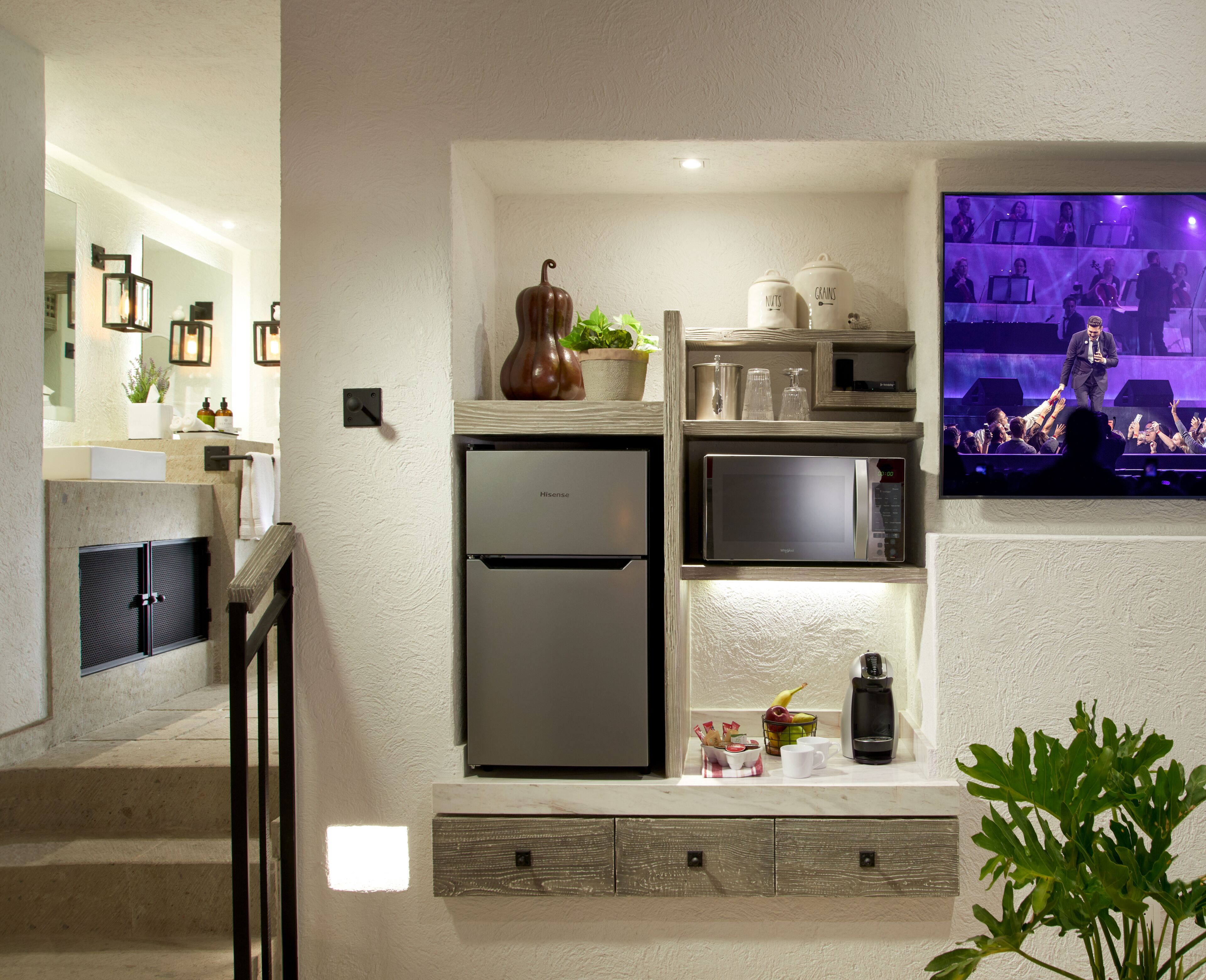 A modern kitchen with a Hisense refrigerator, a microwave, and a coffee maker. There is a small plant on the counter and a TV mounted on the wall displaying a concert.