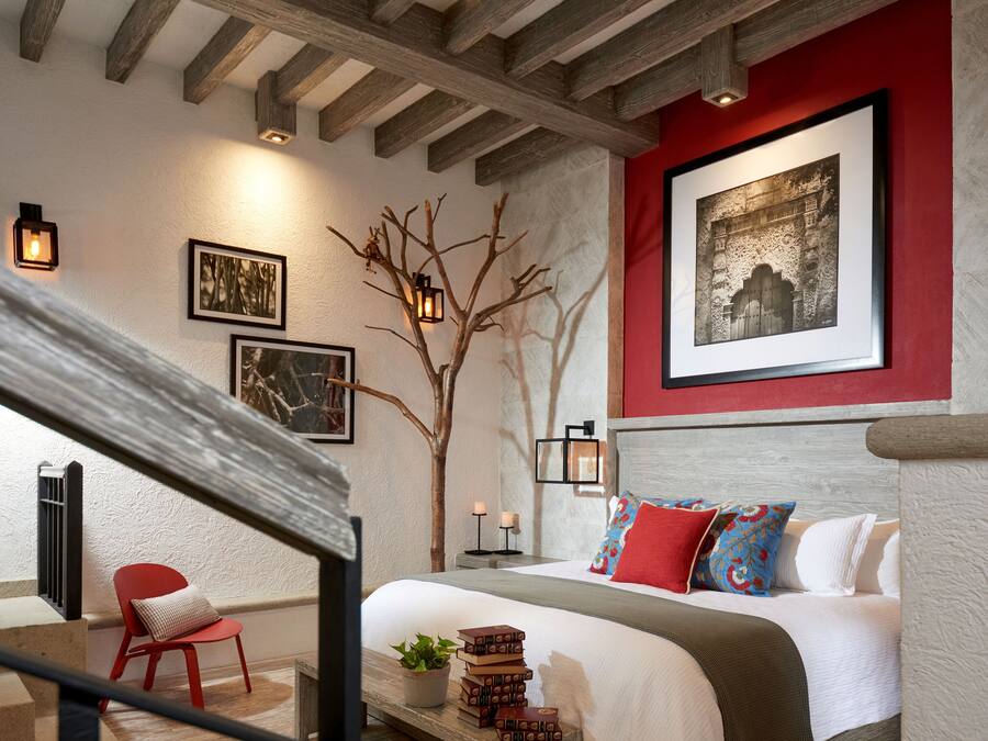 A bedroom with a bed, a red chair, a wooden bench, a potted plant, and a wall-mounted picture.