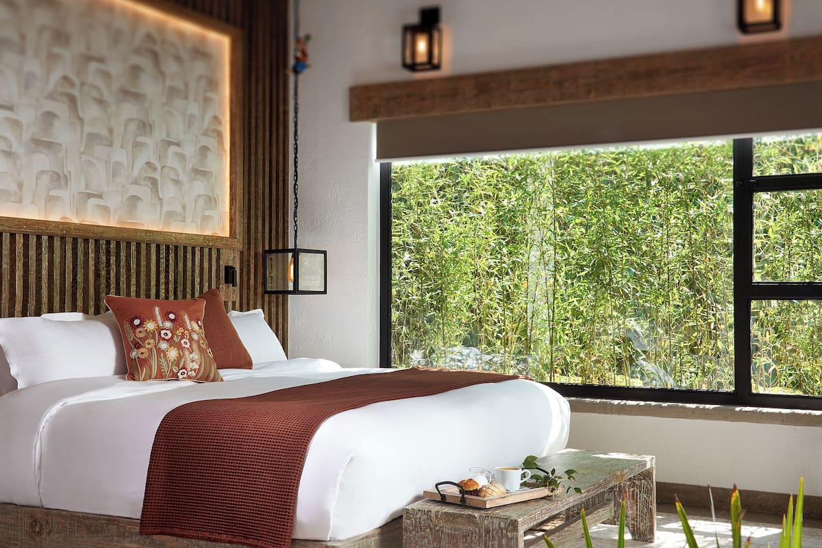 A modern bedroom with a large bed, a wooden headboard, a bedside table with a tray of breakfast items, and a view of greenery through a large window.