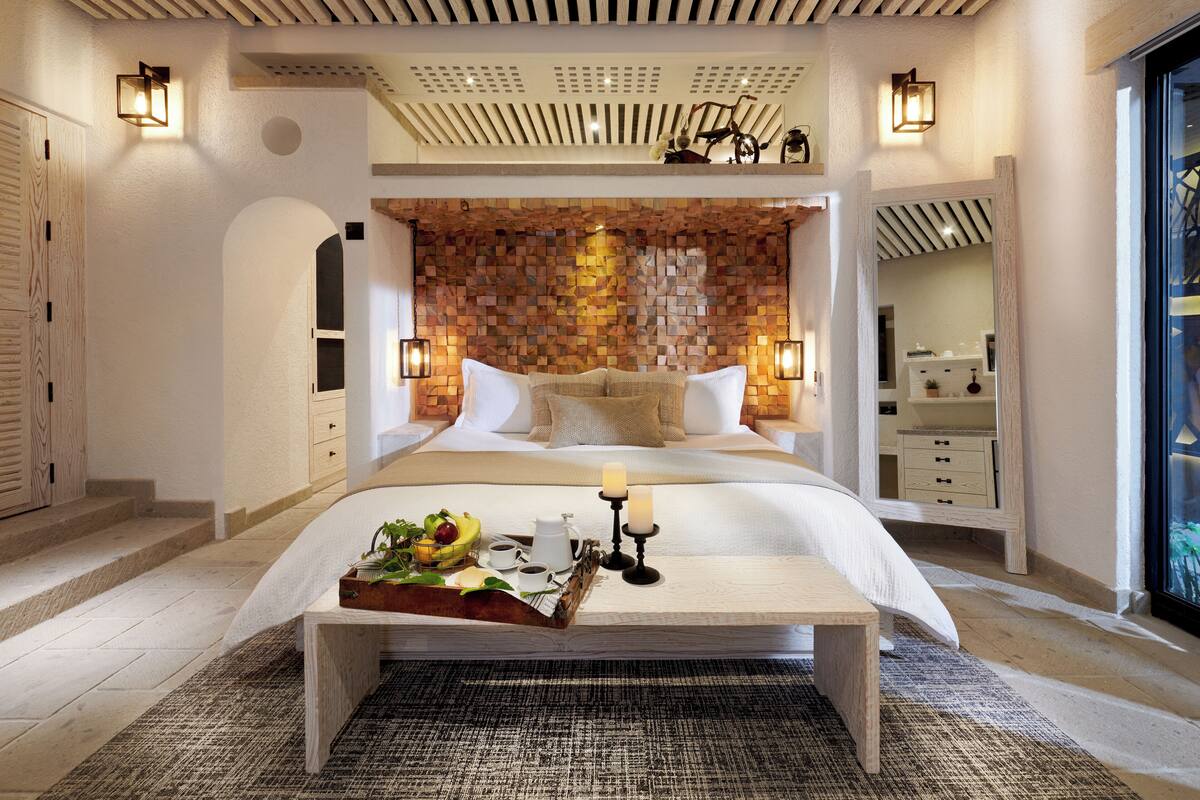 A spacious bedroom with a large bed, a wooden headboard, and a bedside table with a tray of fruits and candles.