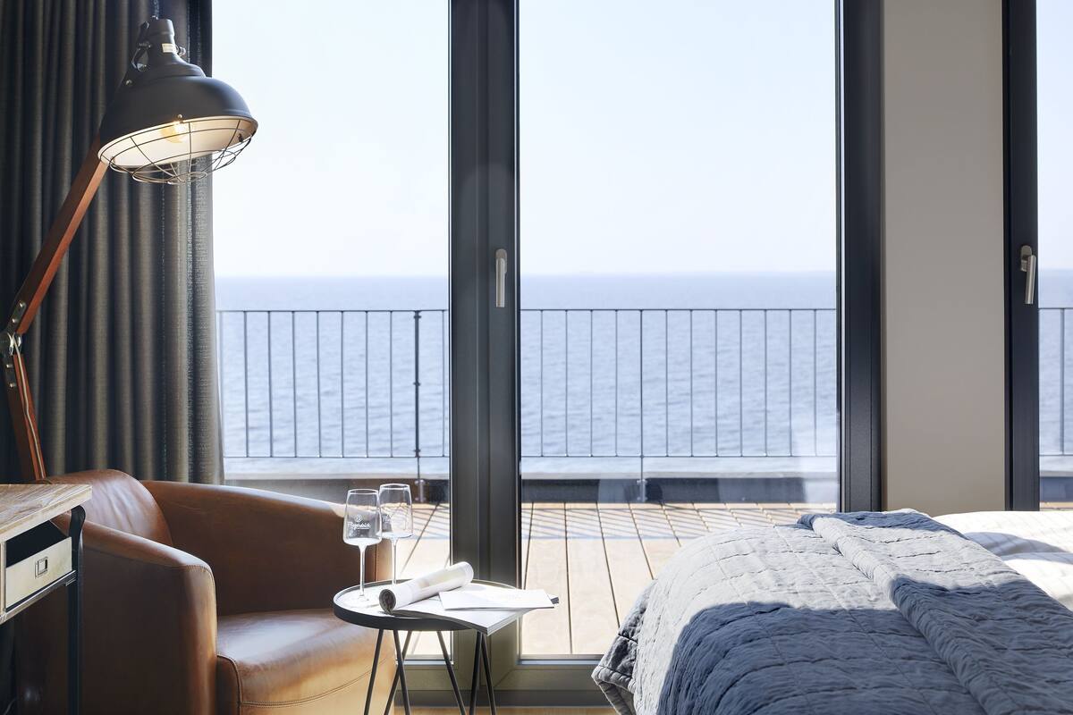 A hotel room with a bed, a desk with a lamp, a chair, a small table with a glass of water and a rolled-up paper, and a balcony with a view of the sea.