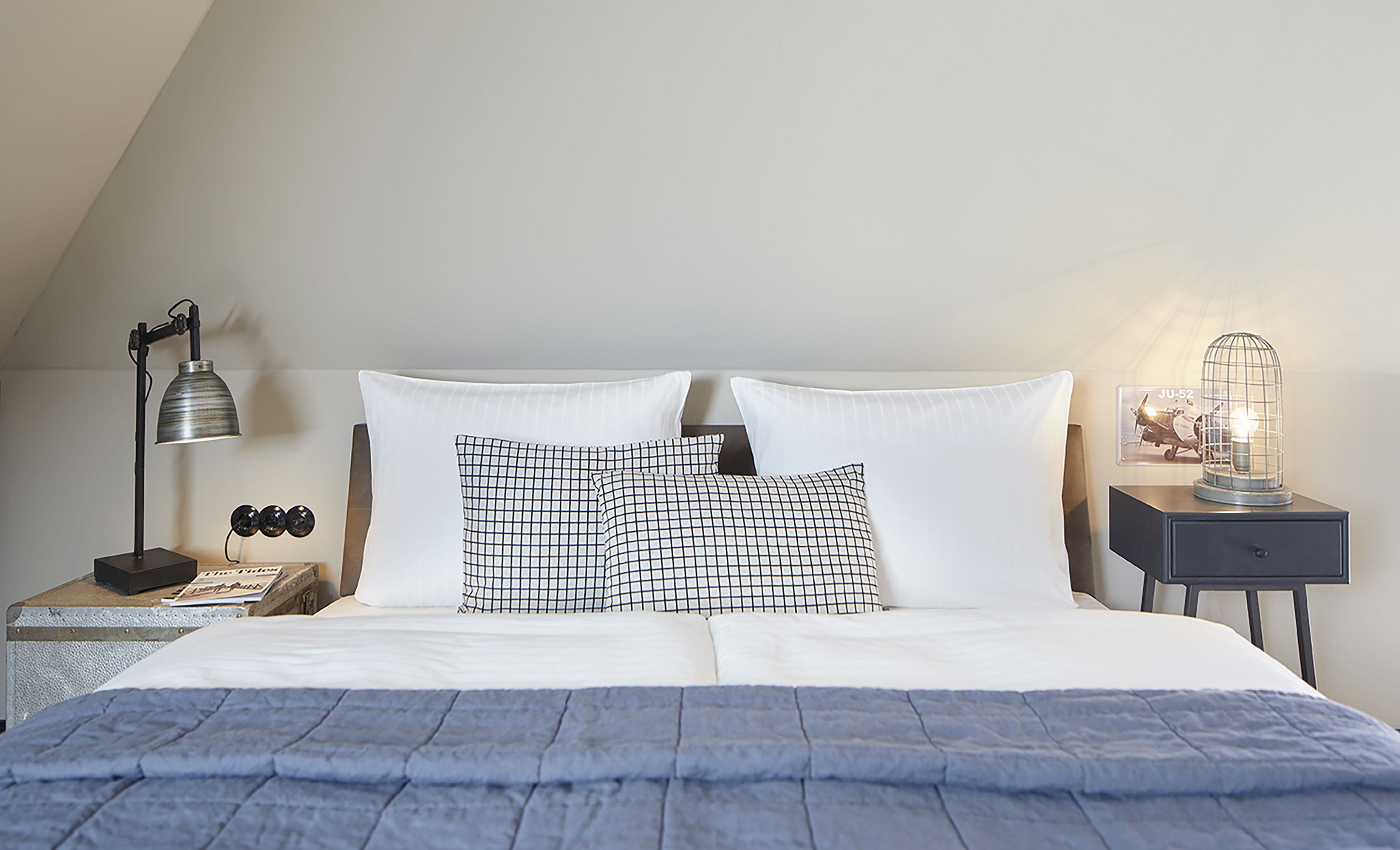 A neatly made bed with white and black checkered pillows, a blue blanket, and a nightstand with a lamp and a vintage light fixture.