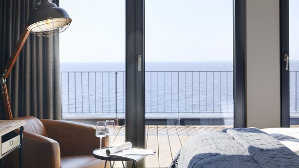 A hotel room with a large window, a bed, a table with a lamp, and a view of the sea. - Hotel Fliegerdeich (Wilhelmshaven)