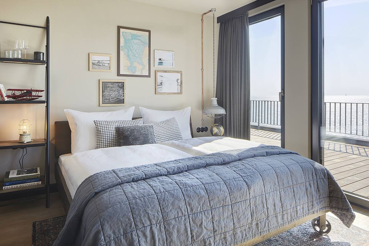 A hotel room with a large bed, a nightstand with books, a map on the wall, and a balcony with a view.