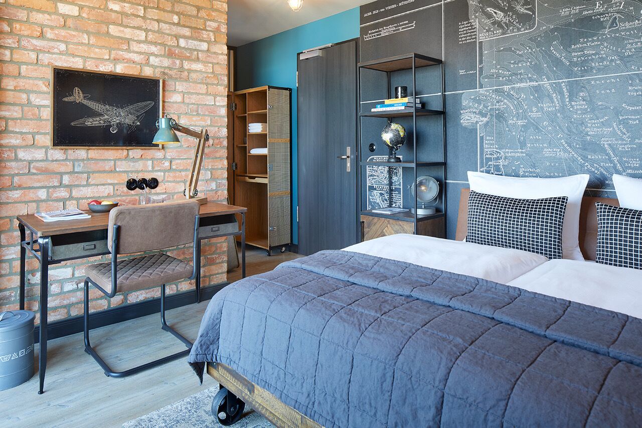 A bedroom with a brick wall, a desk with a chair, a bed with a blue quilt, and a map on the wall.