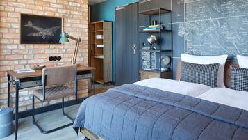 A bedroom with a brick wall, a desk with a chair, a bed with a blue quilt, and a map on the wall.