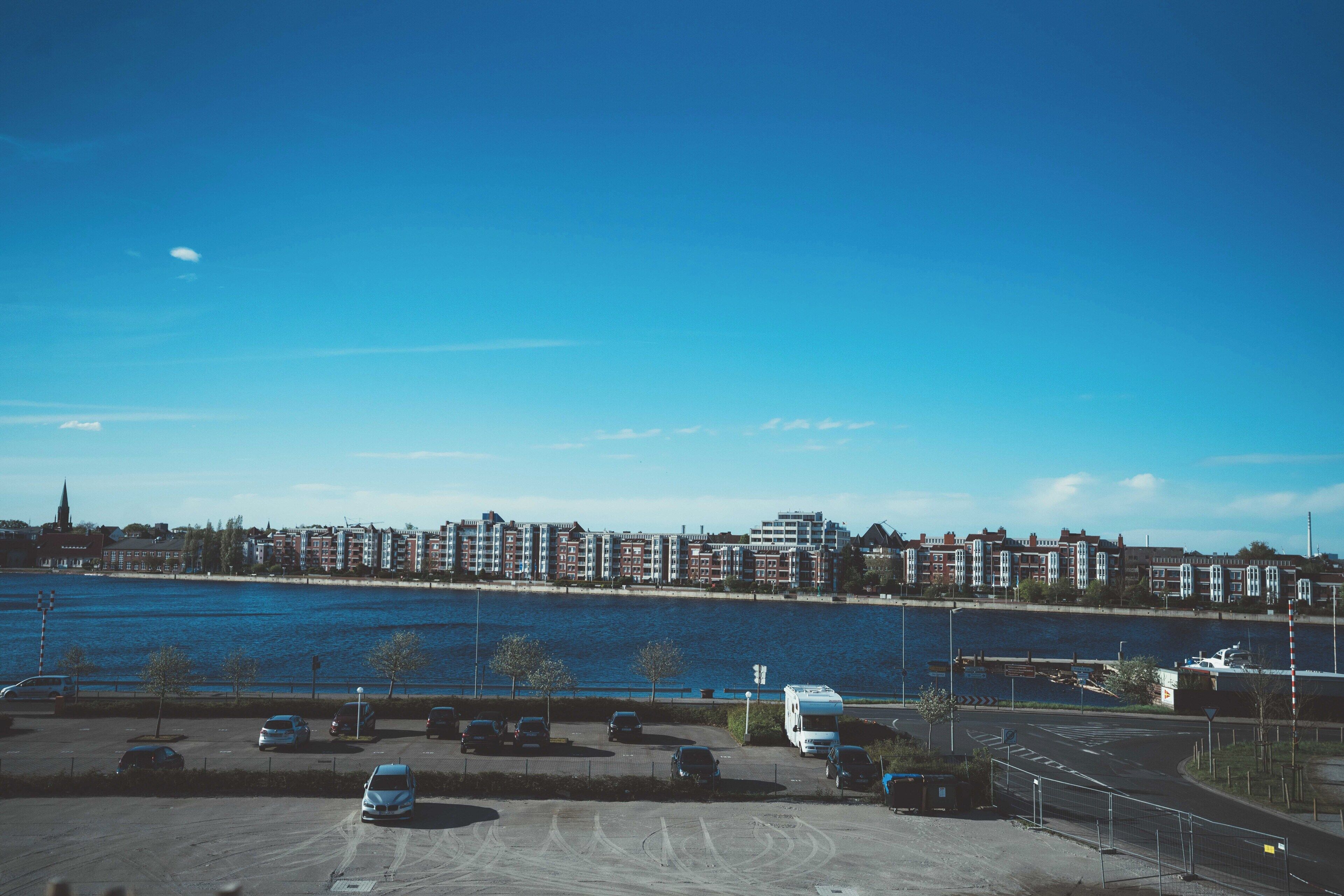 Ein Parkplatz mit mehreren Autos und einem Bus, Blick auf das Wasser und eine Reihe von Gebäuden im Hintergrund.