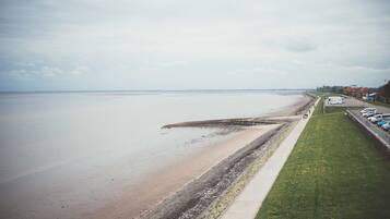 A coastal view with a sandy beach, a grassy promenade, and parked cars along the side.