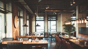 A restaurant with wooden tables and booths, pendant lighting, and large windows allowing natural light. - Hotel Fliegerdeich (Wilhelmshaven)