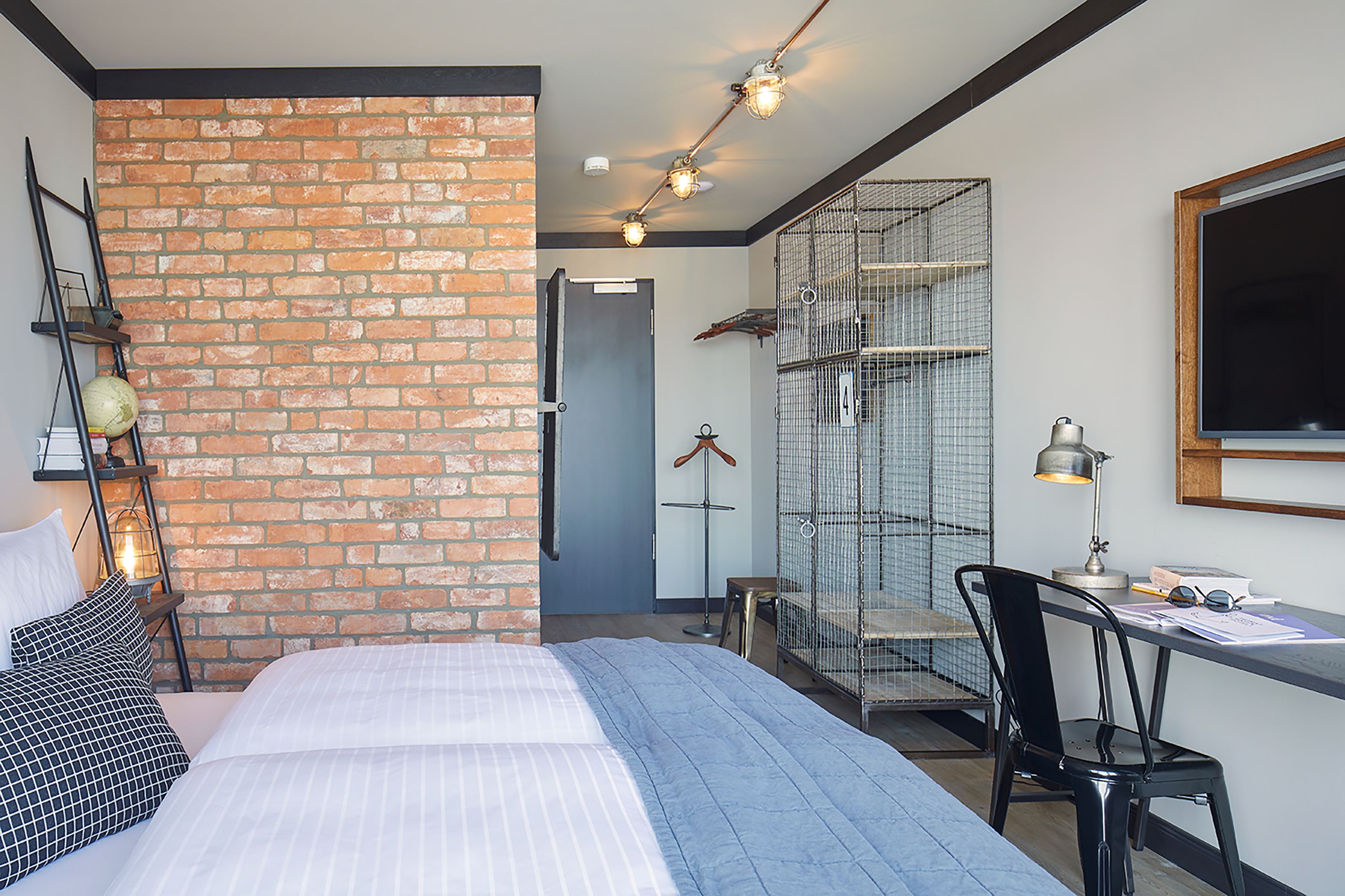 A bedroom with a brick wall, a bed with striped bedding, a desk with a chair, and a hanging light fixture.