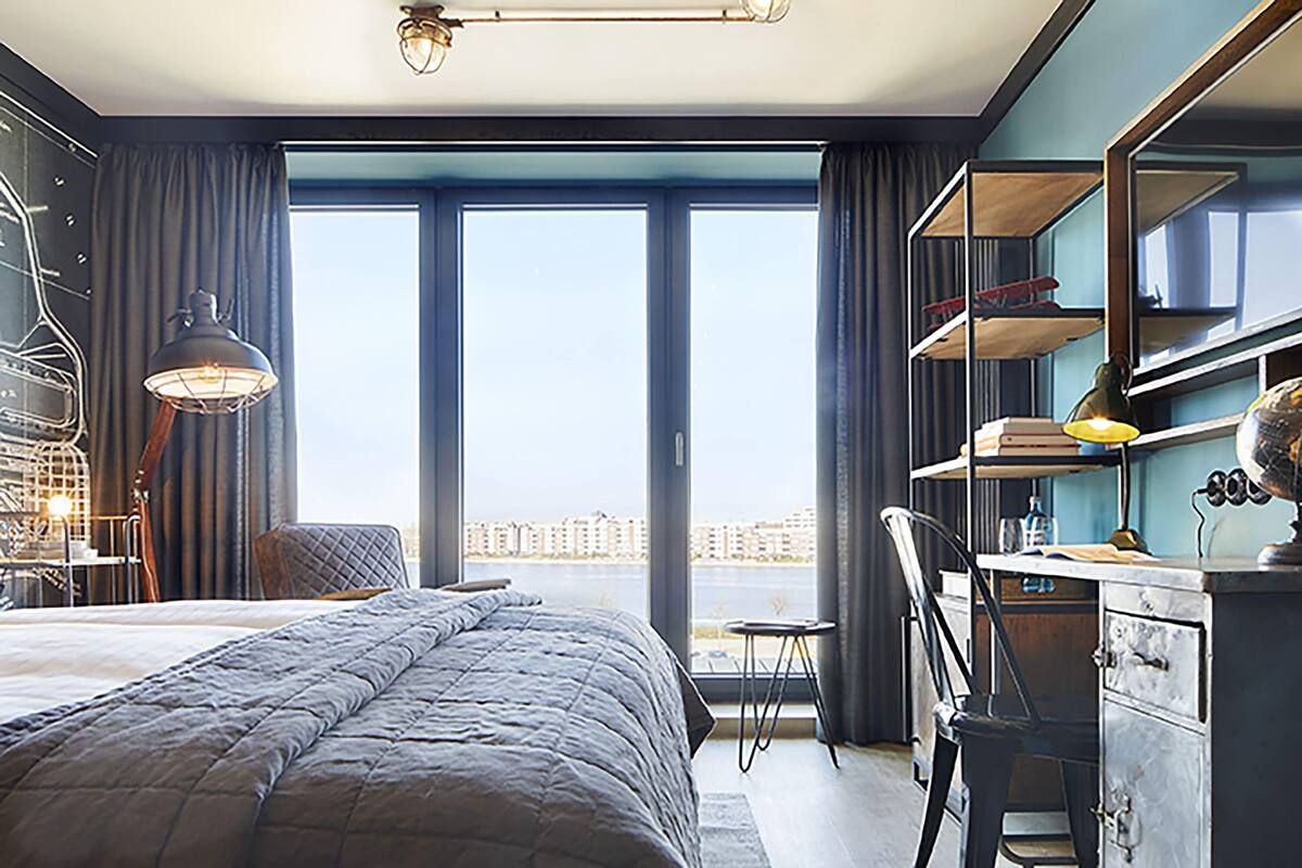 A hotel room with a large bed, a desk, a chair, a desk lamp, a globe, and a view of the city through the window.