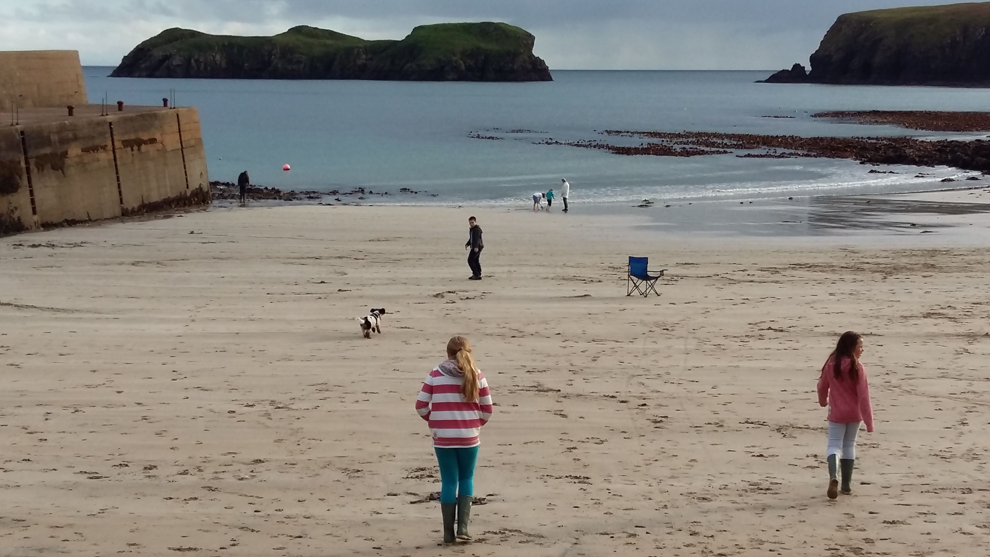 A beach with a concrete barrier, a blue chair, and people walking.