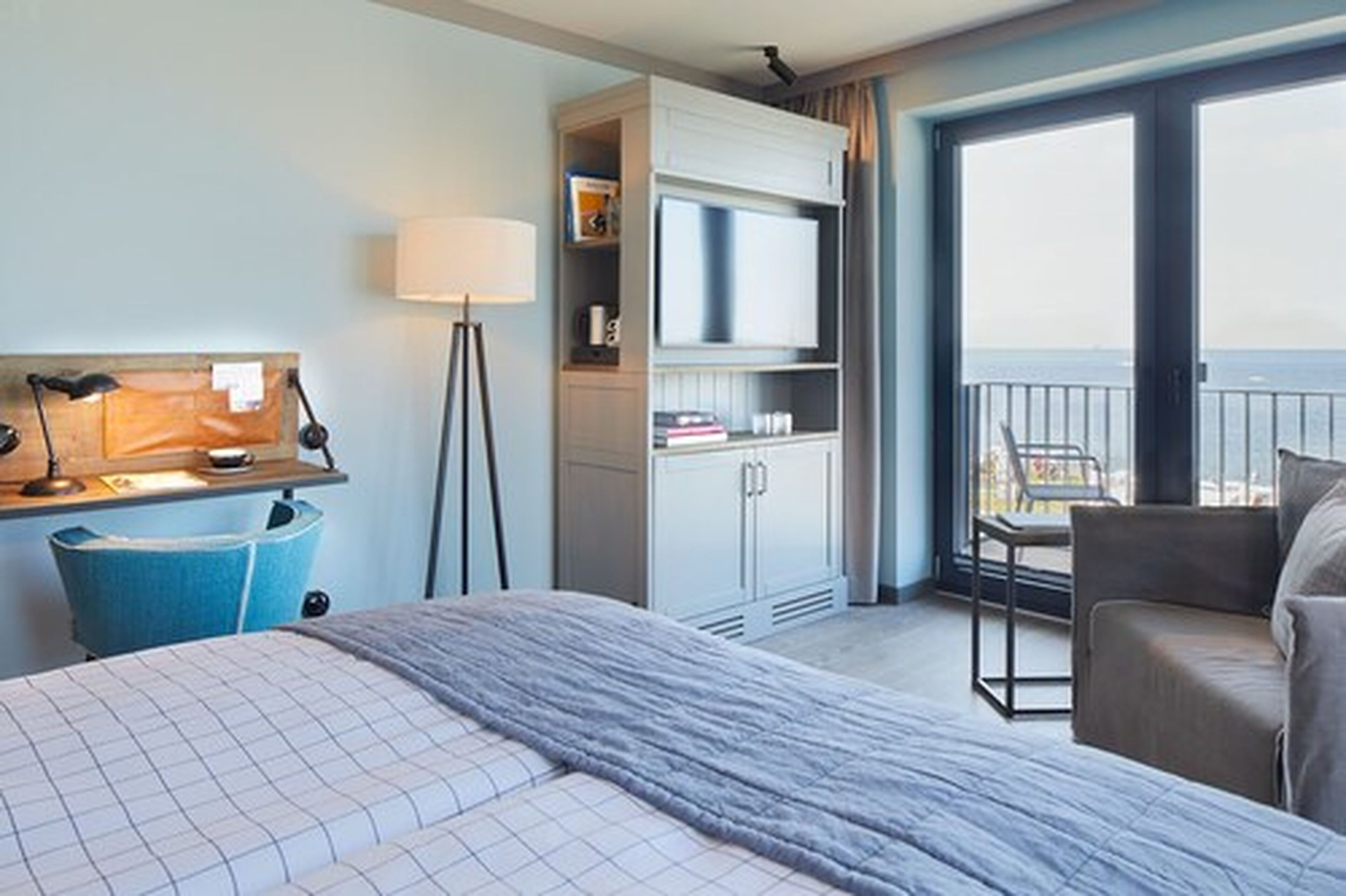 A hotel room with a bed, a desk with a lamp, a TV, a bookshelf, and a balcony with a chair.