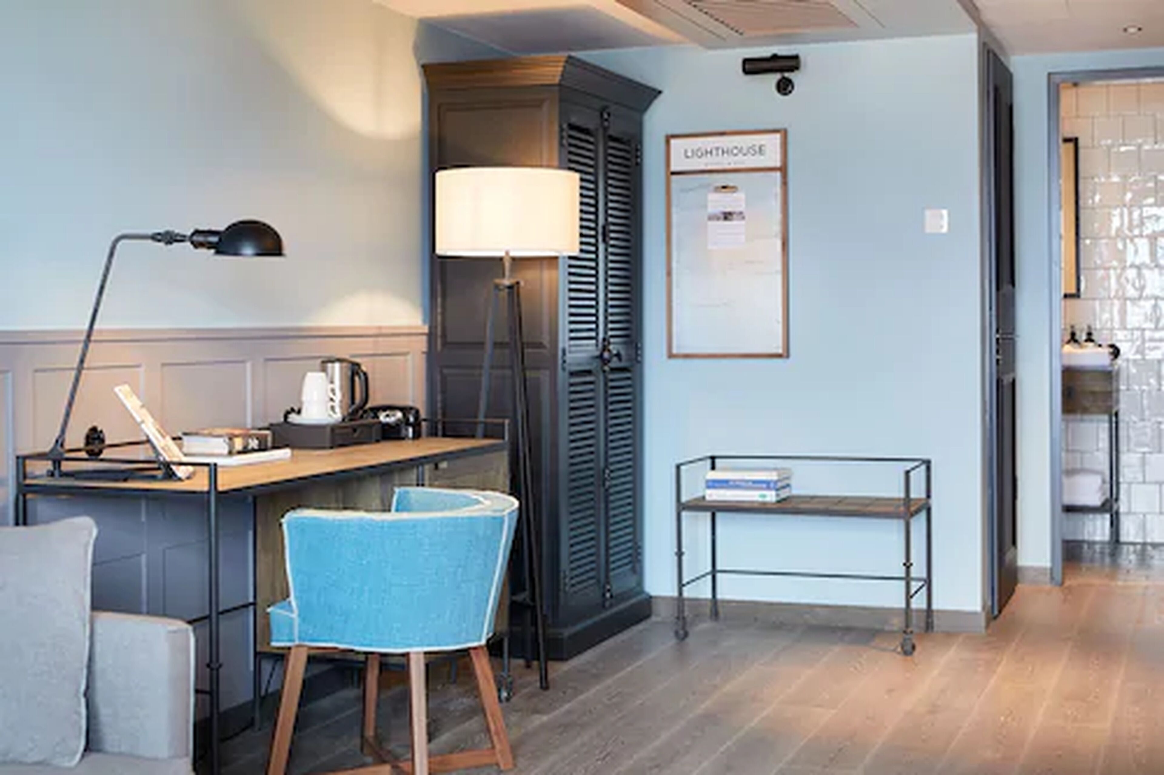 A hotel room with a desk, chair, lamp, and a framed sign on the wall.