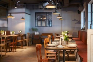 A restaurant with wooden tables and chairs, pendant lighting, and a view of the kitchen through a window.