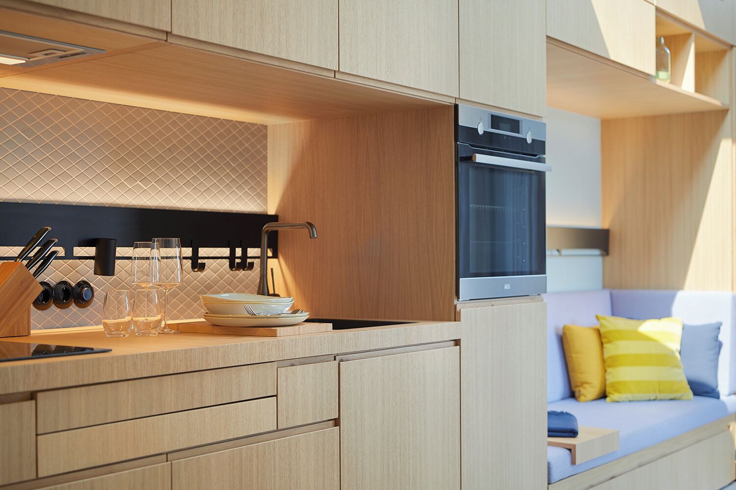 A modern kitchen with wooden cabinetry, a built-in oven, and a dining area with a sofa and cushions.