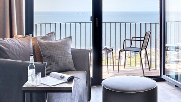A modern living room with a sofa, a small table with a bottle and a glass, and a balcony with a chair. - Lighthouse Hotel & SPA (Büsum)